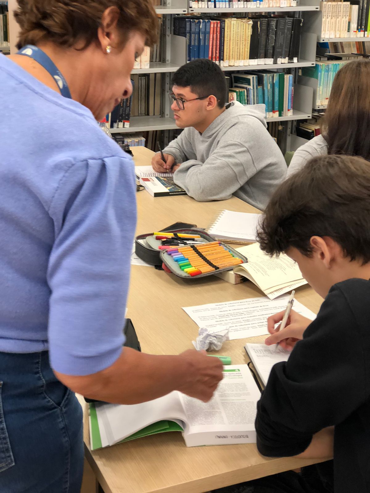 Foto de dois alunos estudando e uma professora ajudando-os com a lição.